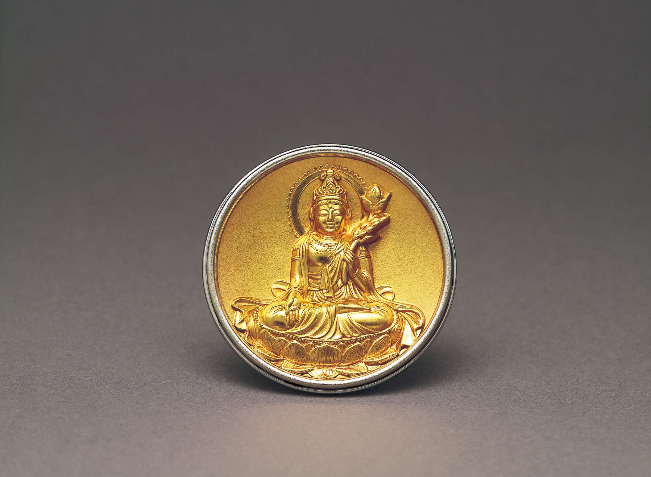 A circular disc of gold depicting in relief a two-armed figure seated on a lotus, holding a stem with a lotus on the end in its left hand, framed by a silver rim.