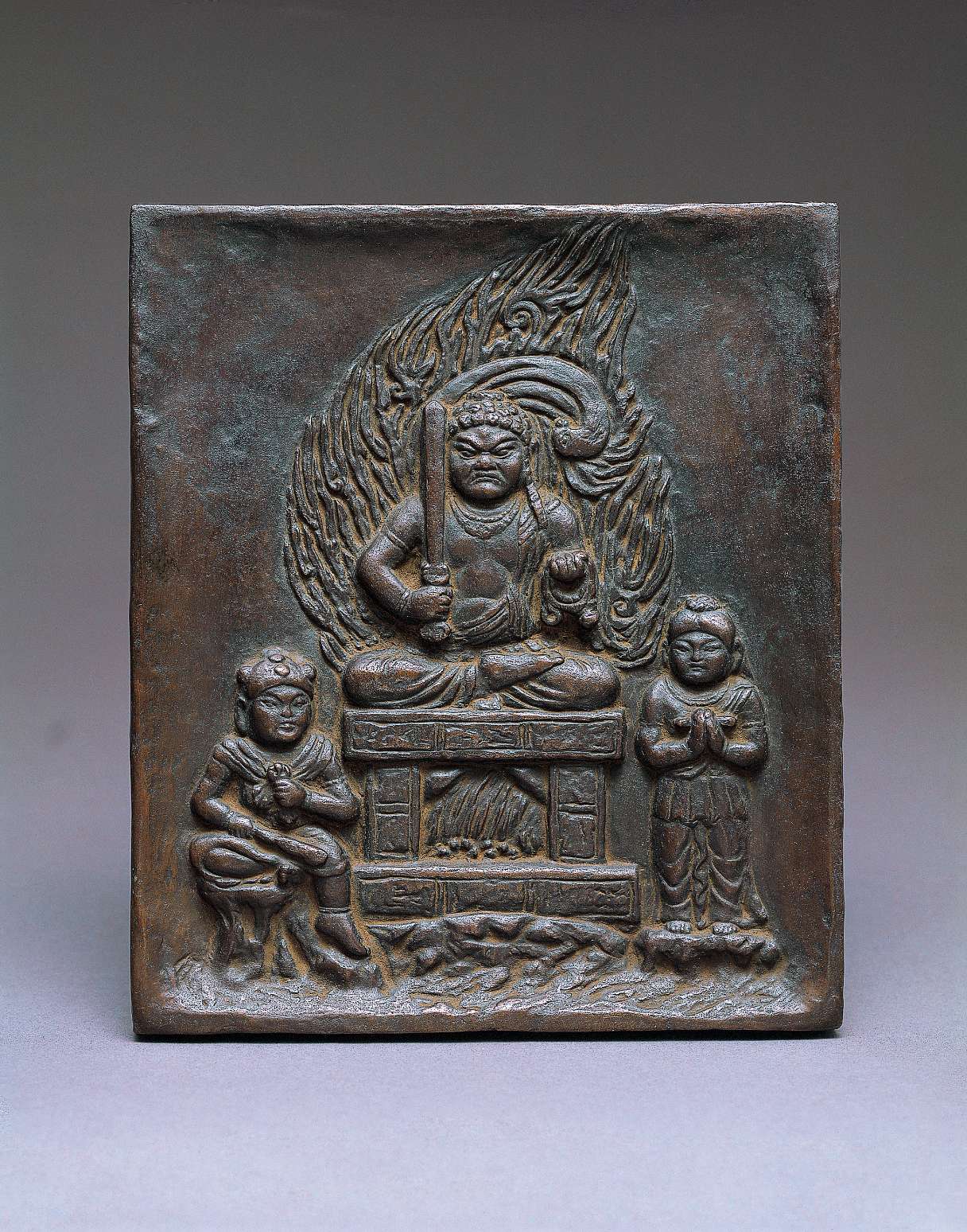 A dark, metallic relief of a scowling figure holding a sword in his right hand and a noose in the left, seated cross-legged on a throne amid a nimbus of stylized fire; two attendants stand to the left and right.