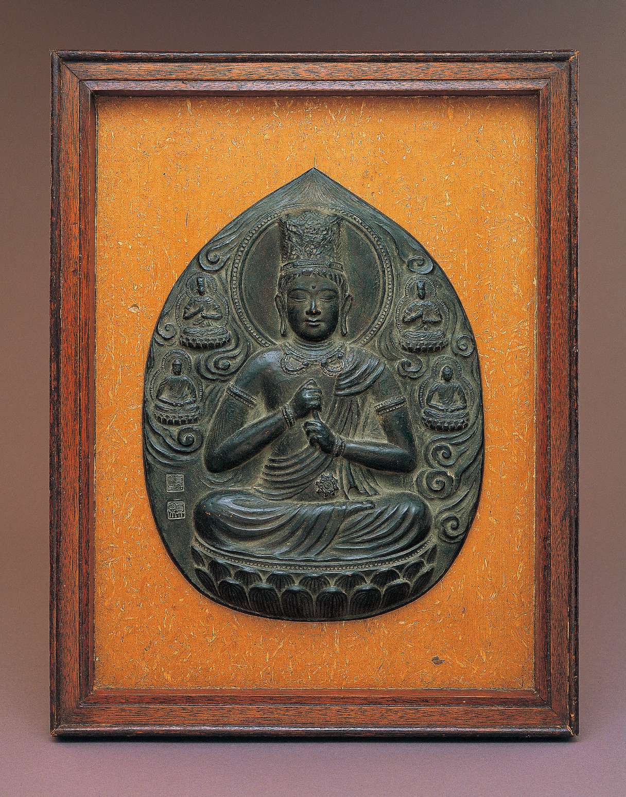An upright almond-shaped metallic relief of a buddha wearing a tall crown sitting cross-legged, grasping his upturned left index finger with his right hand, surrounded by clouds and four smaller buddhas.