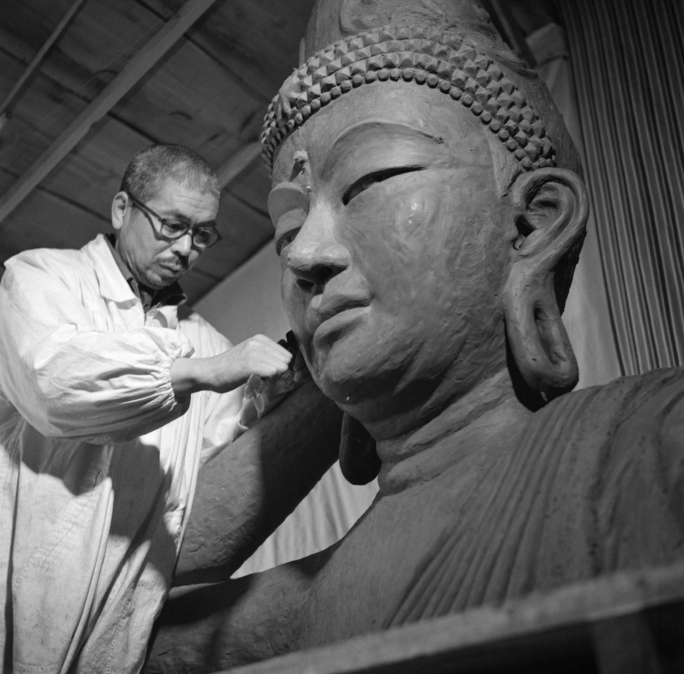 Shinjo, with sculpting implements in hand, wearing a smock over his clothing, stands sculpting a very large bust of a smiling buddha.