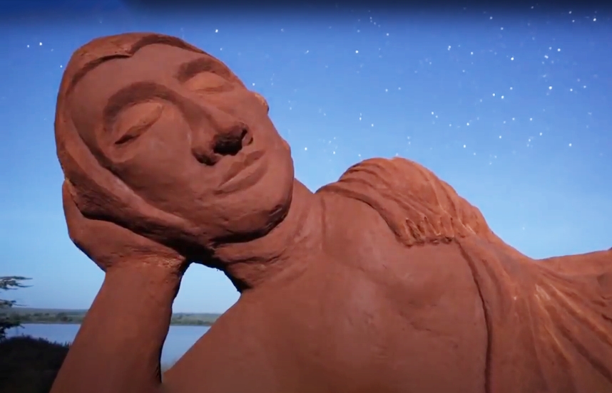 A large earthen sculpture of the Buddha laying on his right side, head supported by right arm, is visible against a dusk sky in which stars are visible.