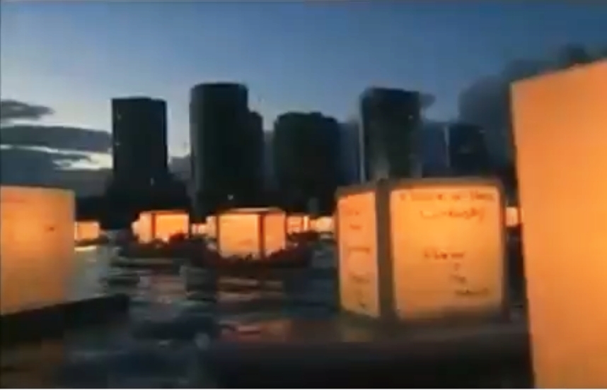 Lanterns illuminated by candles within float in a bay with a cityscape and evening sky visible in the background.