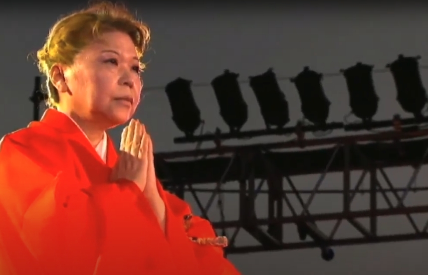Her Holiness, in bright orange robes, presses her palms together in prayer beneath an outdoor evening sky.