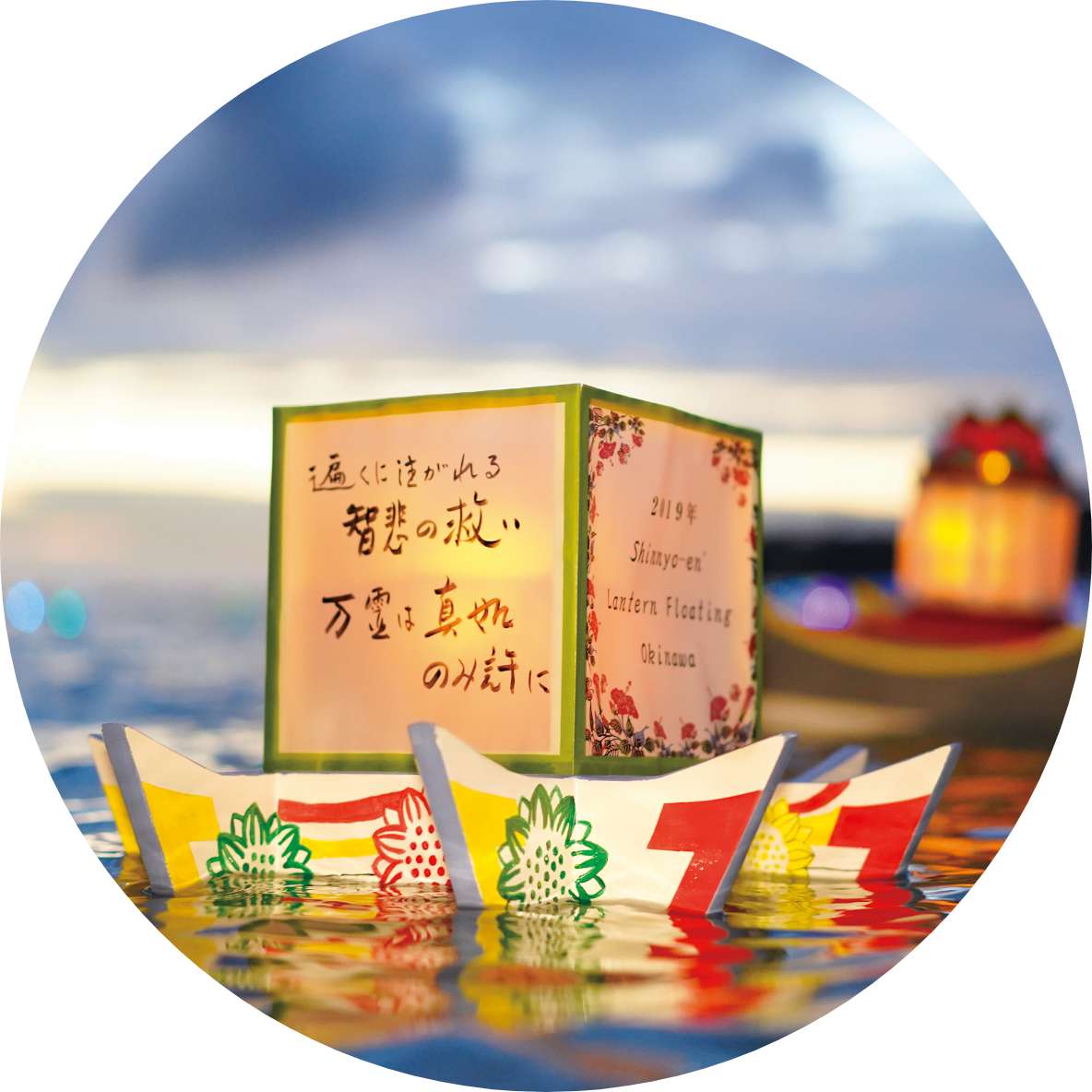 An illuminated paper lantern, hand decorated with messages in Japanese, floats on a colorful base, with the evening sky in the background.