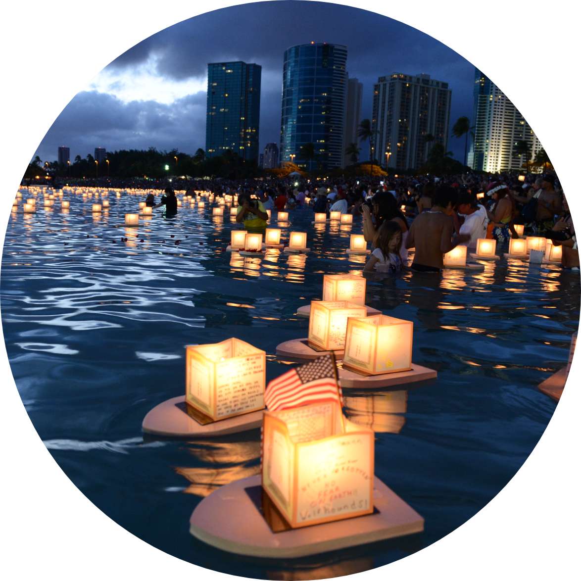 Glowing lanterns float in calm waters of an ocean bay with a high-rise cityscape in the background.